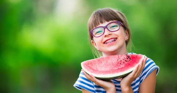 Child eating watermelon. Kids eat fruits in the garden. Pre teen girl in the garden holding a slice of water melon. happy girl kid eating watermelon. Girl kid with gasses and teeth braces — стоковое фото