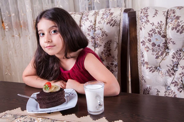 Молодой красивый маленький ребенок Ближнего Востока девушка с Шоколадный торт с ананасом, клубникой и молоком с красным платьем и темными глазами и длинными волосами счастливым пить и есть дома и сидит улыбается. глядя на камеру. студия выстрел — стоковое фото