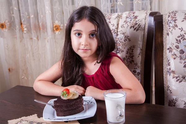 Молодой красивый маленький ребенок Ближнего Востока девушка с Шоколадный торт с ананасом, клубникой и молоком с красным платьем и темными глазами и длинными волосами счастливым пить и есть дома и сидит улыбается. глядя на камеру. студия выстрел — стоковое фото