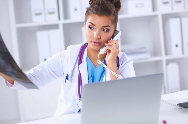 Женский доктор смотреть рентгеновского сканирования и разговаривает по телефону в диагностический центр, сидя за столом — стоковое фото