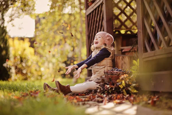 Милая счастливая детская девочка, играющая и бросающая листья в солнечный осенний сад — стоковое фото