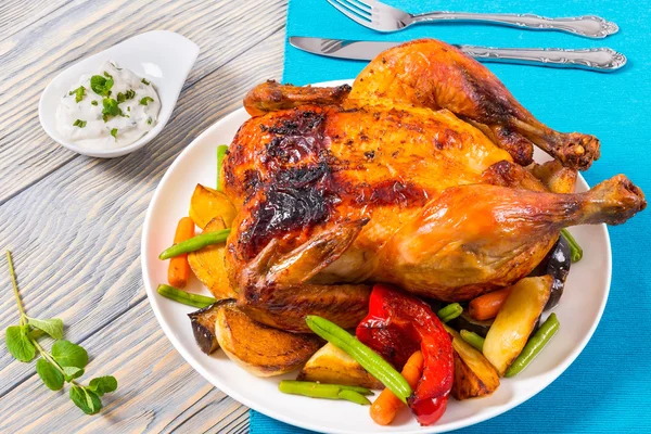 Roasted whole chicken with potatoes, baby carrots, eggplants and green beans on the white round dish on the table napkin, with mint yogurt sauce, Christmas festive dinner, top view — стоковое фото