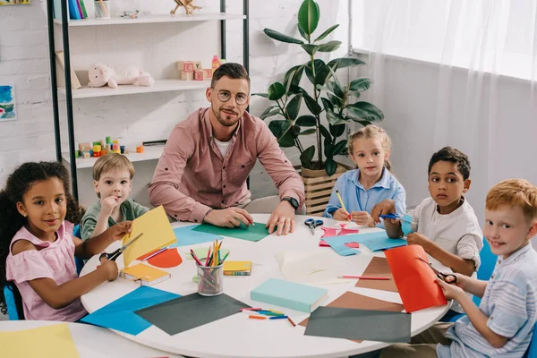 Male Teacher Multiracial Preschoolers Sitting Table Colorful Papers Classroom — стоковое фото