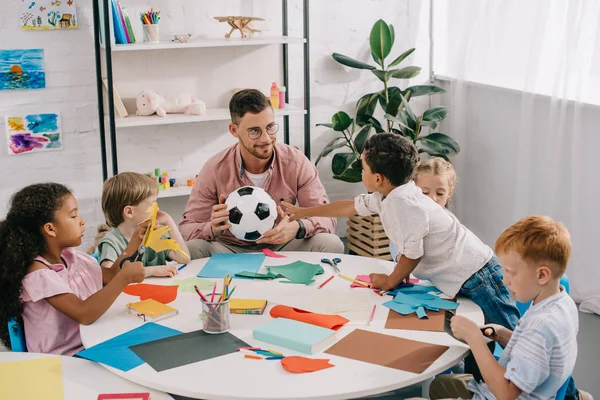 Male Teacher Soccer Ball Multiracial Preschoolers Sitting Table Colorful Papers — стоковое фото