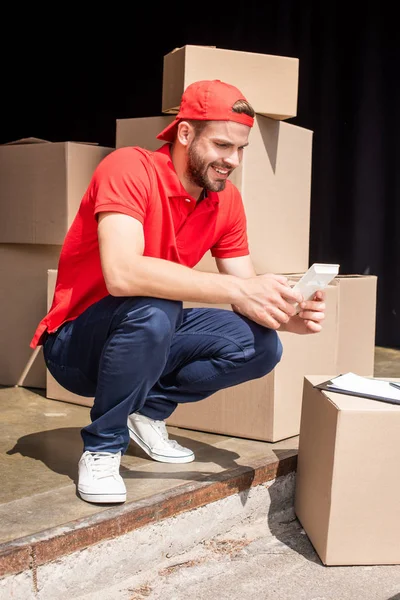Young Smiling Delivery Man Making Calculations Calculator Cardboard Boxes — стоковое фото