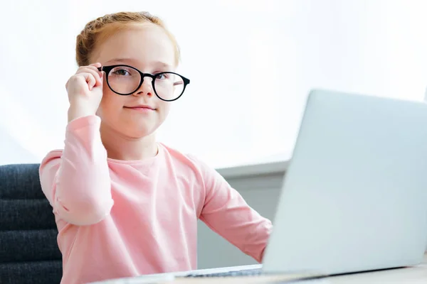 Beautiful Schoolchild Adjusting Eyeglasses Smiling Camera While Using Laptop — стоковое фото