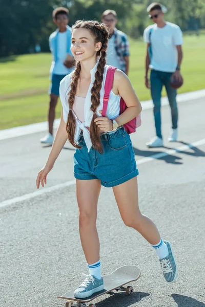 Smiling Teenage Girl Backpack Riding Skateboard While Classmates Walking Park — стоковое фото