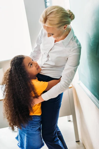 Adorable African American Schoolgirl Embracing Her Teacher Classroom — стоковое фото